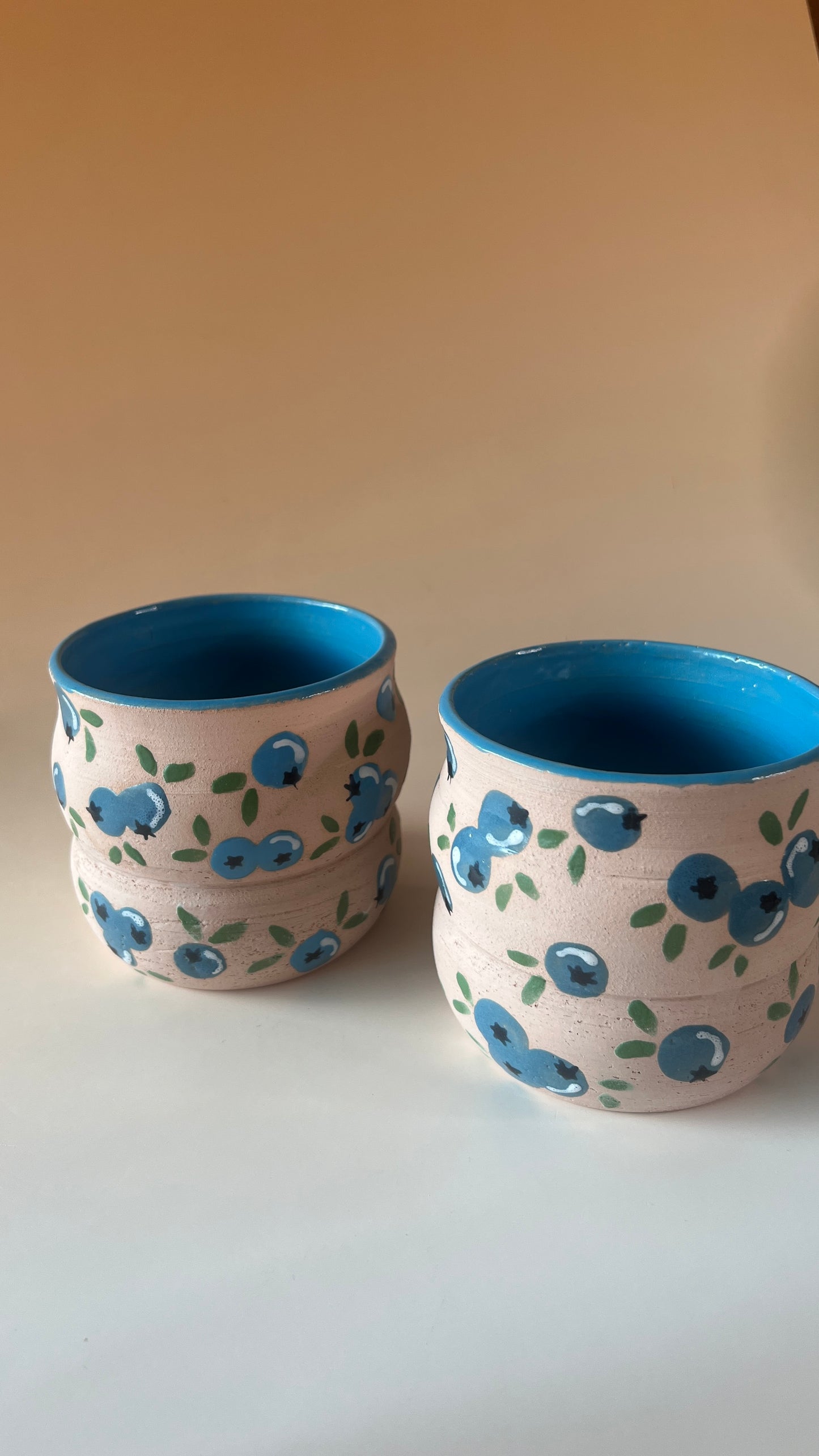 Two ceramic cups with blue interior and blueberries exterior design on a beige background