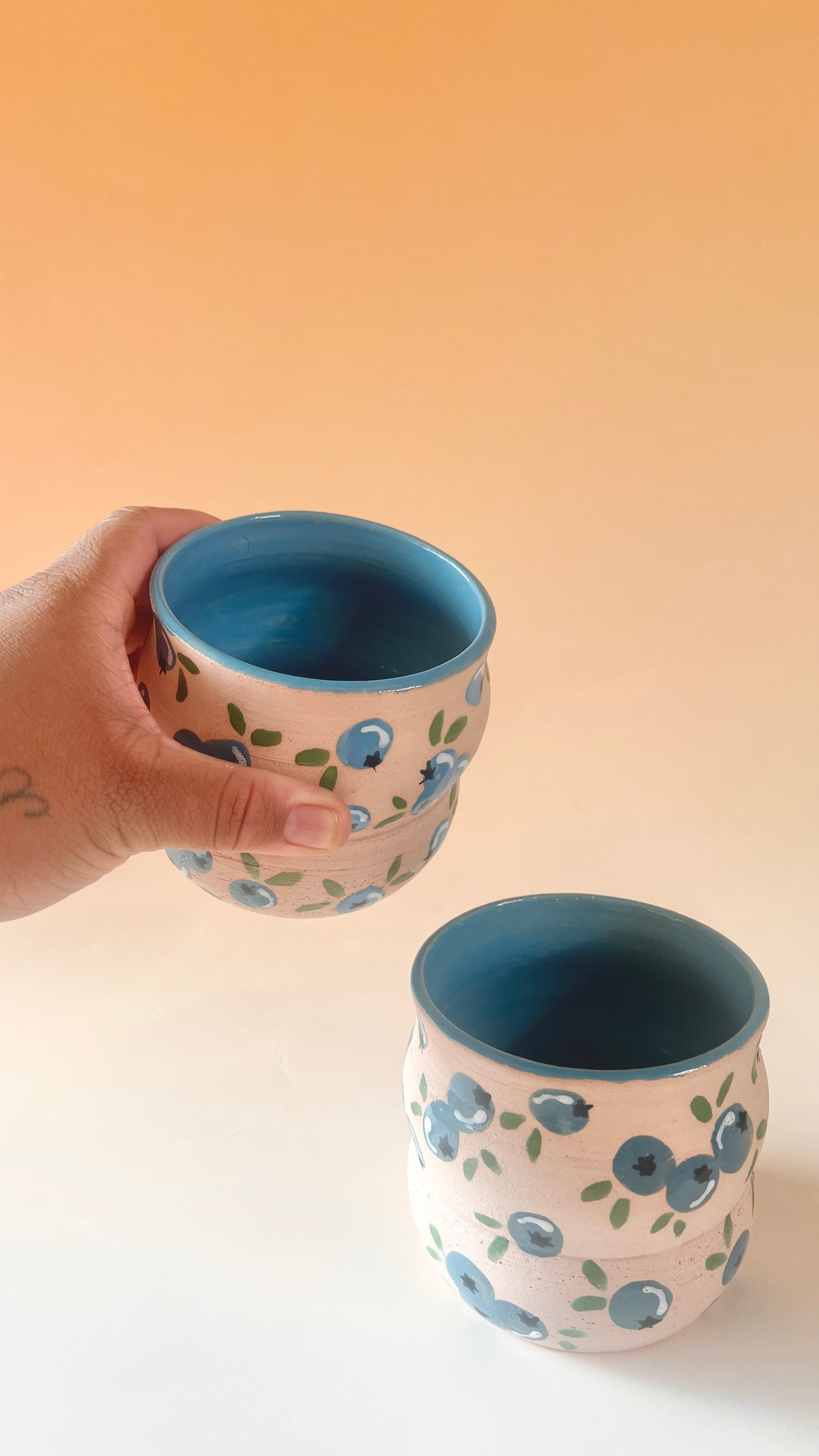 Two ceramic cups with blue interior and blueberry exterior design, one held by a hand against a beige background.