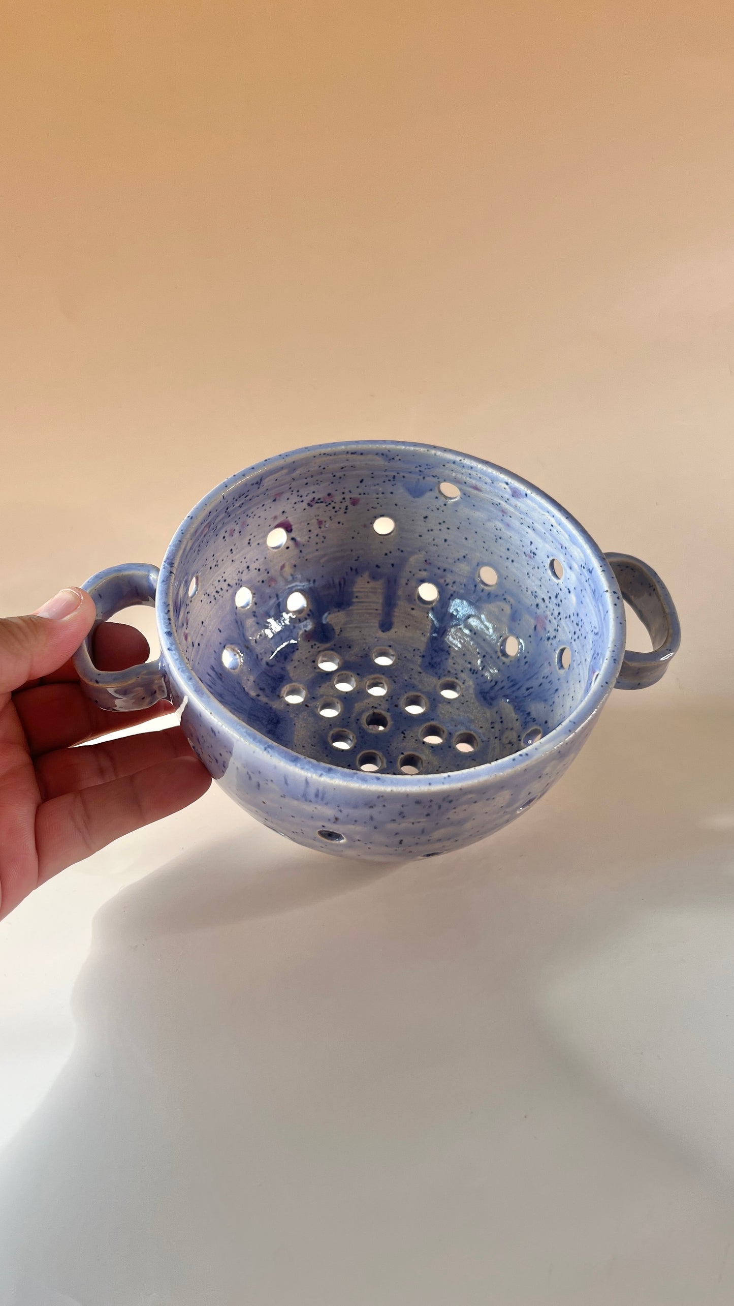 Hand holding a small ceramic colander with a speckled blue pattern on a beige background