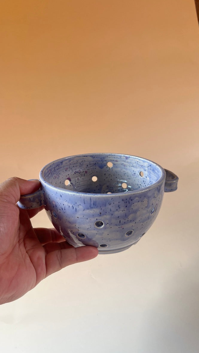Hand holding a blue ceramic pot with holes against a plain background