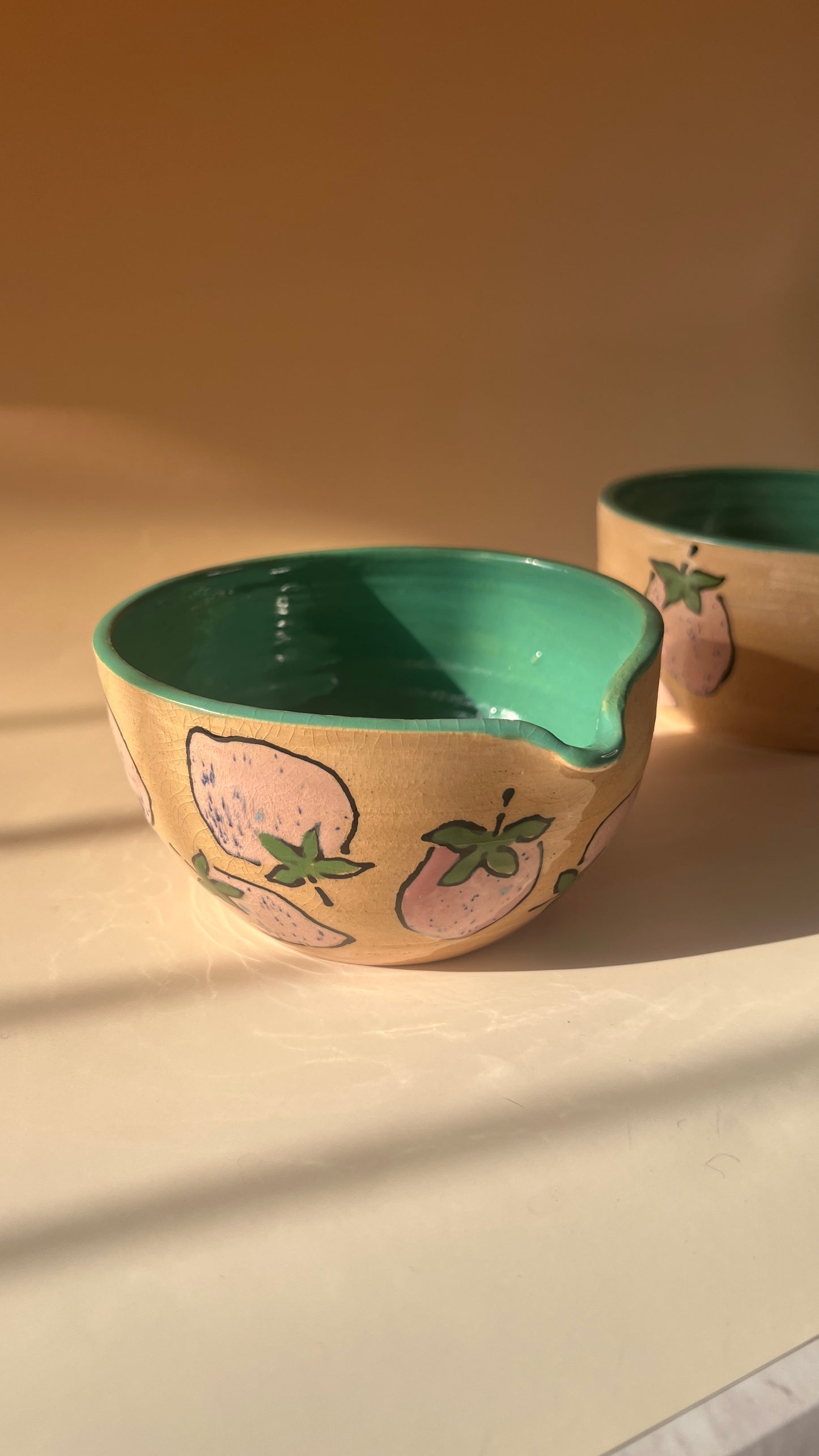 Ceramic bowl with green interior and pink exterior featuring a spout and pink strawberries on a beige background