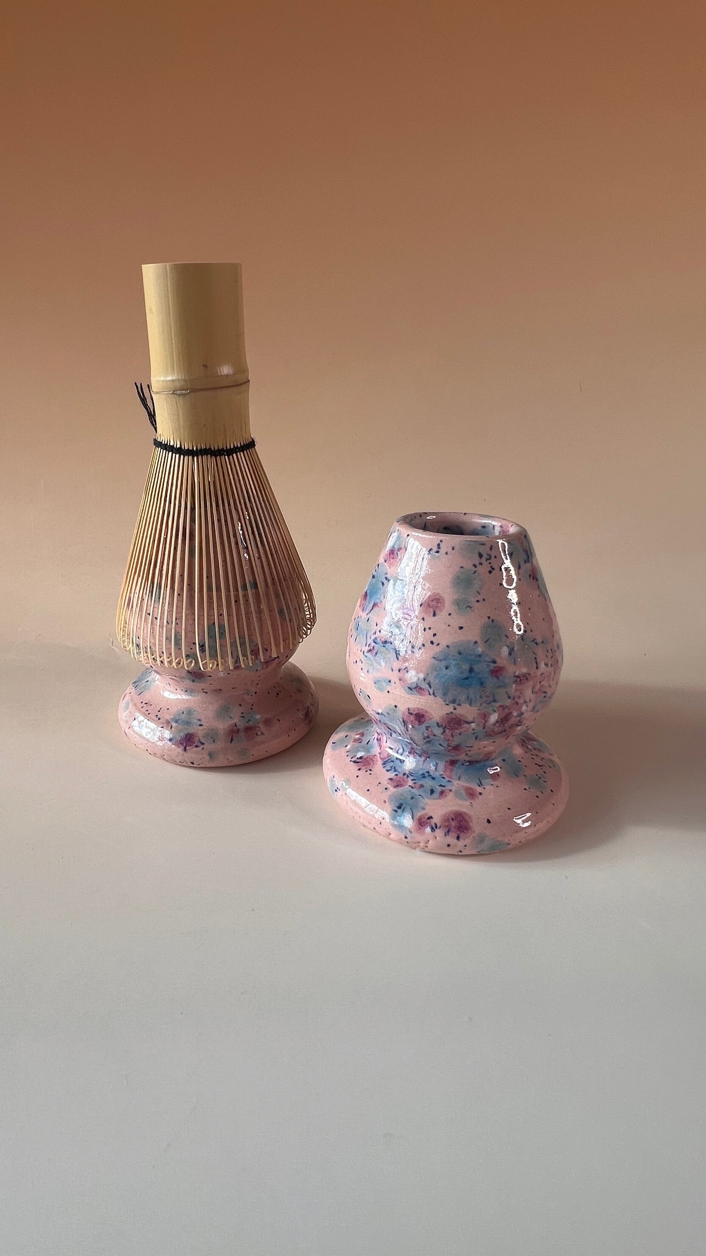 Two pink ceramic matcha whisk holders with colorful blue, pink, and white speckles on a beige backgroundwith colorful blue, pink, and white speckles on a beige background