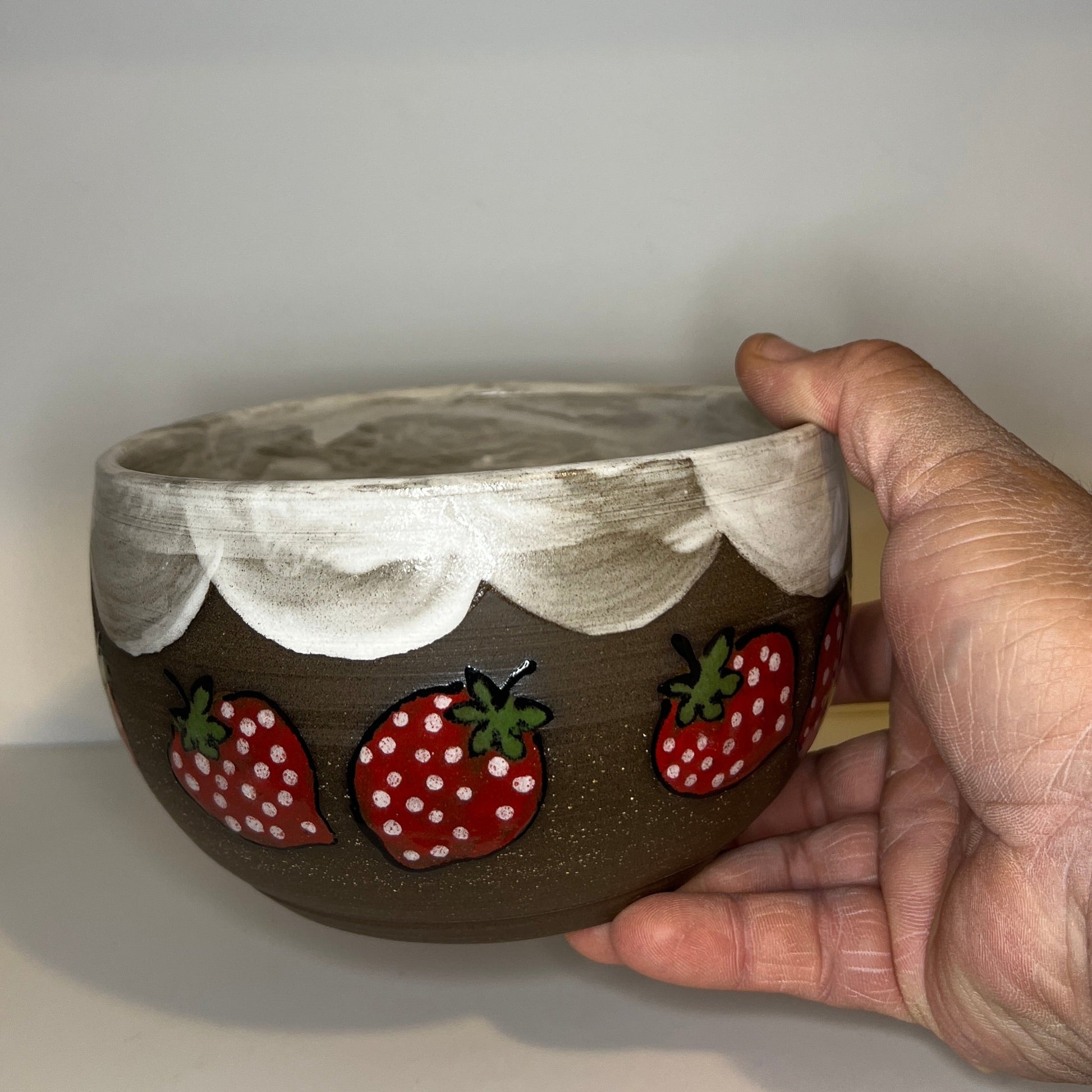 Dark brown ceramic bowl with strawberry designs and added with a red inside and The red rim features a hand-glazed swooping design that adds movement and visual interest. The bowl is against a white background and held by a hand 
