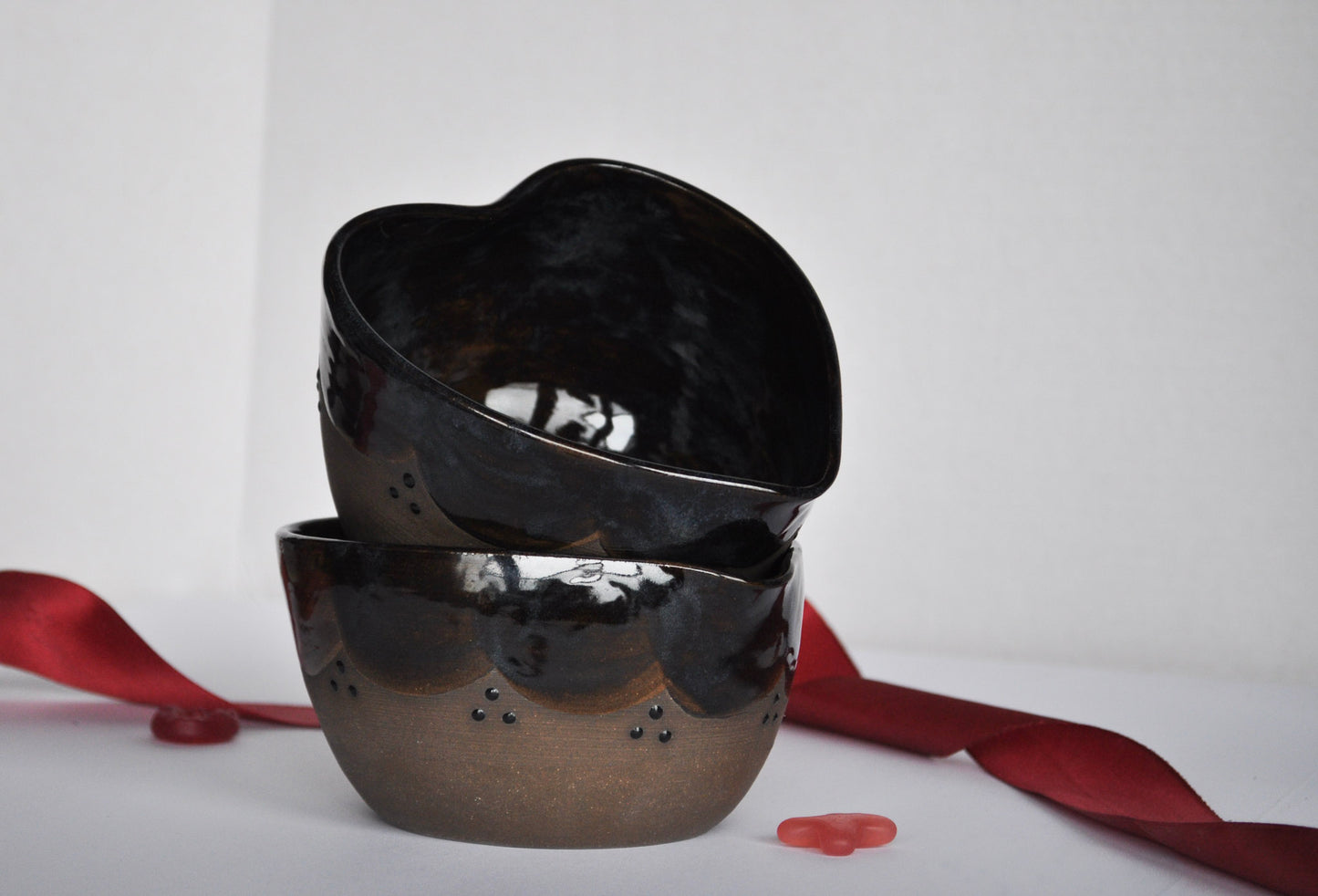 Two black heart-shaped bowls stacked on a white surface with red ribbons and petals.