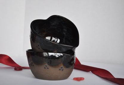 Two black heart-shaped bowls stacked on a white surface with red ribbons and petals.