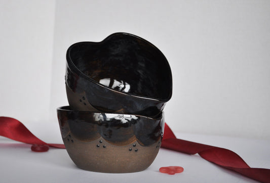 Two black heart-shaped bowls stacked on a white surface with red ribbons and petals.