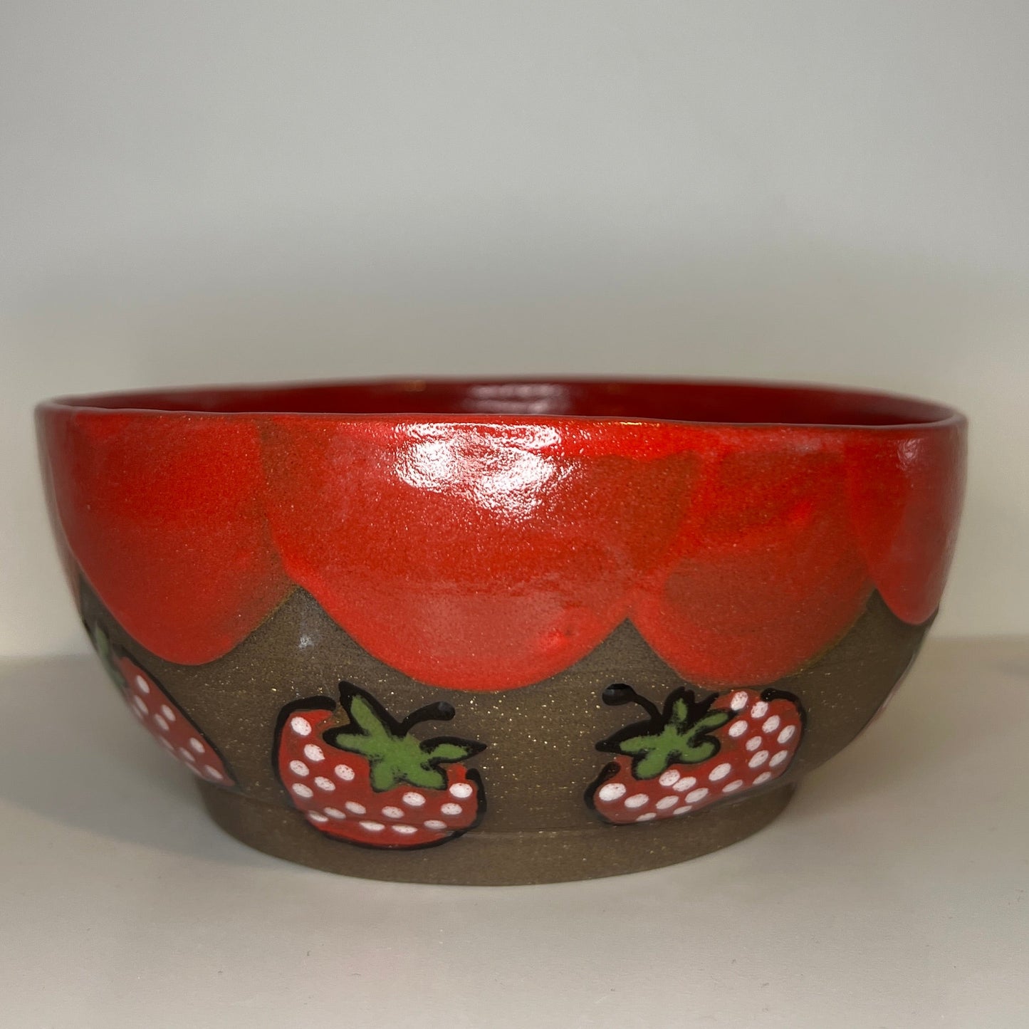 Dark brown ceramic bowl with strawberry designs and added with a red inside and The red rim features a hand-glazed swooping design that adds movement and visual interest. The bowl is against a white background.