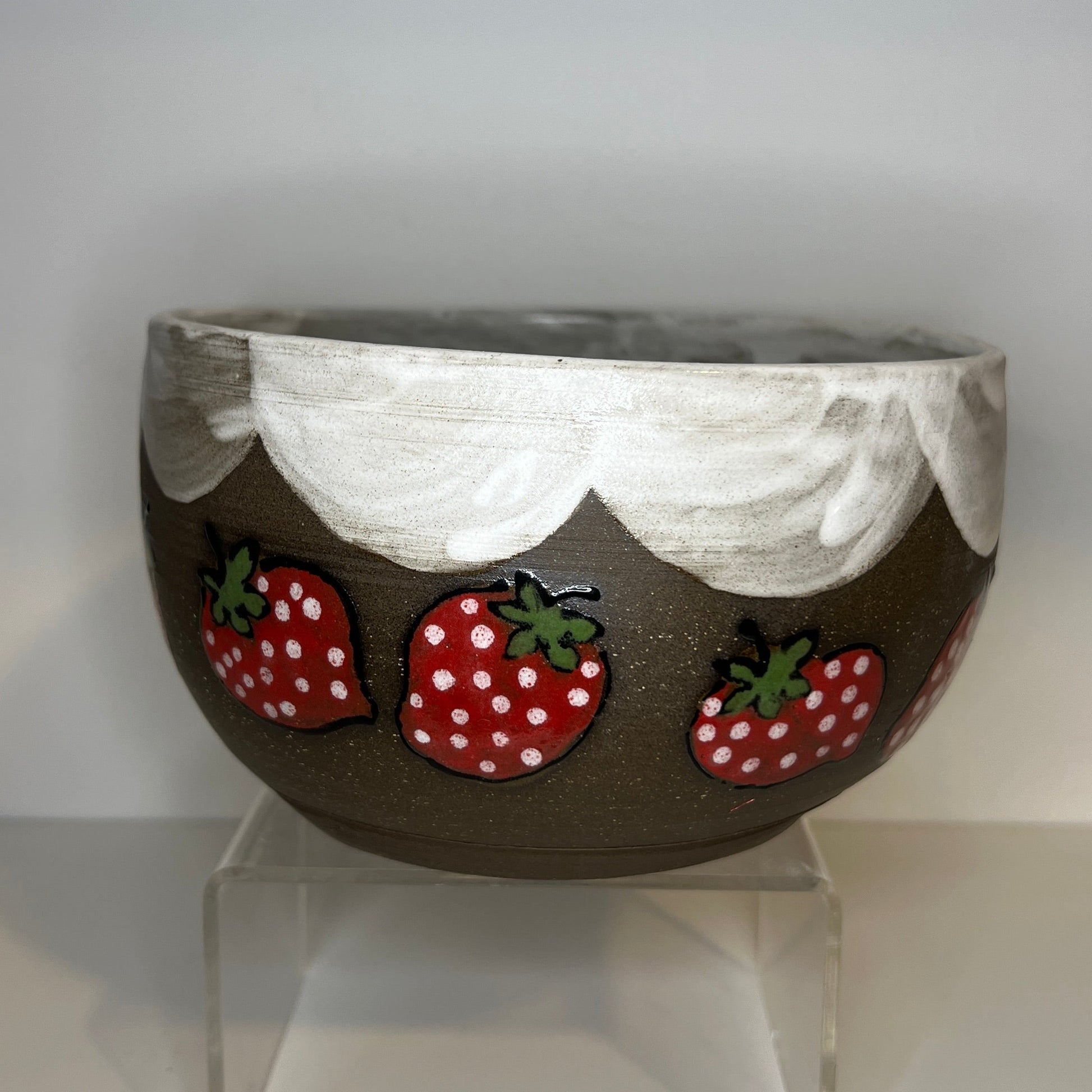Dark brown ceramic bowl with strawberry designs and added with a white color inside and The white rim features a hand-glazed swooping design that adds movement and visual interest. The bowl is on a clear stand against a white background