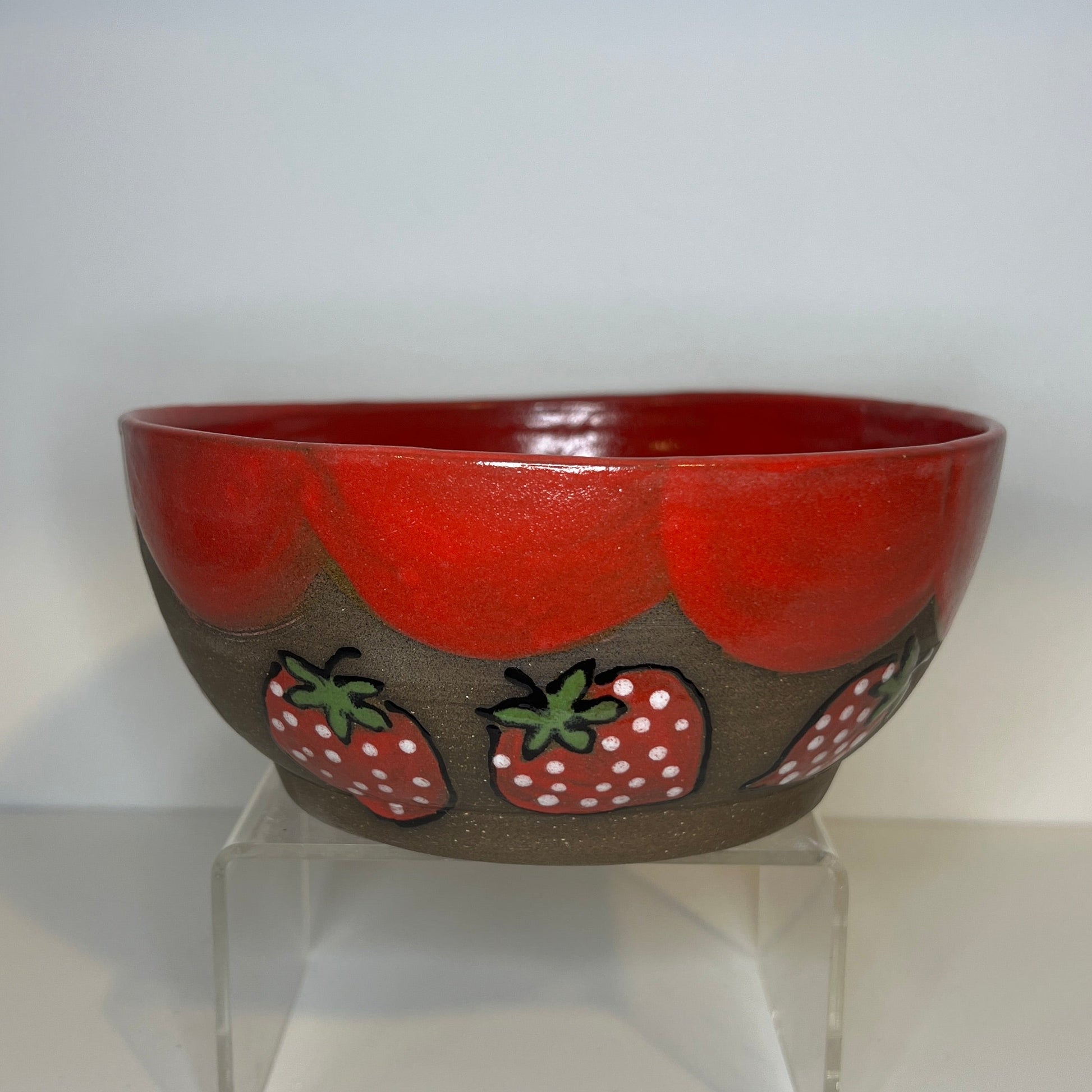 Dark brown ceramic bowl with strawberry designs and added with a red inside and The red rim features a hand-glazed swooping design that adds movement and visual interest. The bowl is on a clear stand against a white background