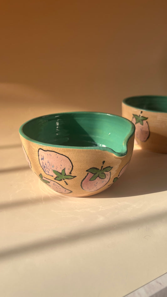 Ceramic bowl with green interior and pink exterior featuring a spout and pink strawberries on a beige background